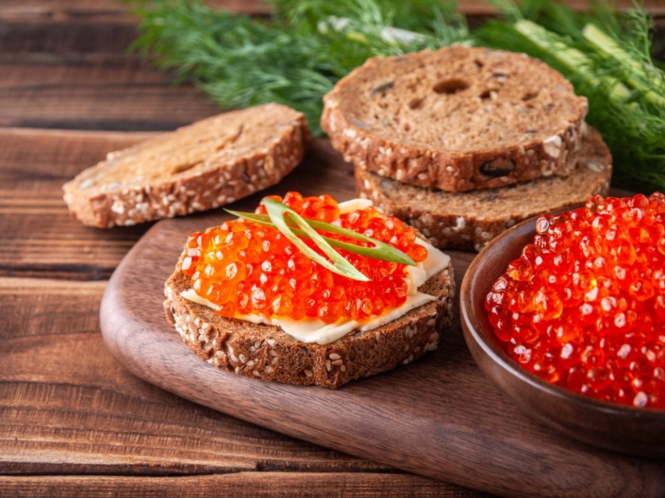 Sandwich with red caviar on a wooden tray surrounded by bread, caviar and herbs